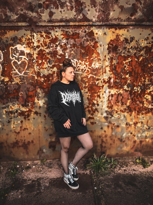 Ganzkörperansicht eines Models im schwarzen Doombound-Hoodie vor einer rostigen Wand